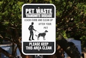 A black and white sign with text warning about pet waste transmitting disease. It instructs pet owners to leash and clean up after their pets. The sign features an illustration of a person walking a dog. It is mounted on a post surrounded by trees.