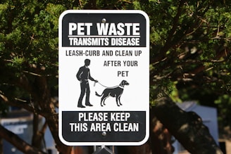 A black and white sign with text warning about pet waste transmitting disease. It instructs pet owners to leash and clean up after their pets. The sign features an illustration of a person walking a dog. It is mounted on a post surrounded by trees.