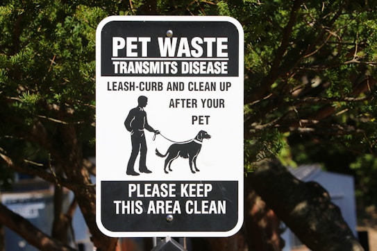 A black and white sign with text warning about pet waste transmitting disease. It instructs pet owners to leash and clean up after their pets. The sign features an illustration of a person walking a dog. It is mounted on a post surrounded by trees.
