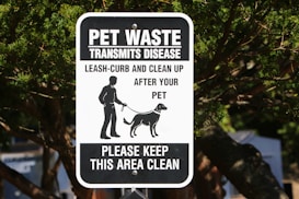 A black and white sign with text warning about pet waste transmitting disease. It instructs pet owners to leash and clean up after their pets. The sign features an illustration of a person walking a dog. It is mounted on a post surrounded by trees.