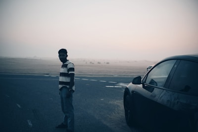 A person stands on a road beside a car, with a vast desert landscape in the background under a hazy sky.