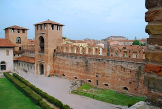A historic brick fortress with tall towers and crenellated walls stands prominently. The fortress has multiple levels, with an archway entrance and a courtyard area below. Surrounding buildings with red-tiled roofs are visible in the background, and well-manicured green hedges and grass are present in the foreground.