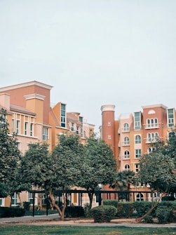 A cluster of multi-story residential buildings with an architectural style reminiscent of Mediterranean or European influences. The buildings feature arched windows and balconies, with a color palette of warm earth tones like peach and terracotta. In the foreground, there are several lush, green trees and neatly trimmed bushes, suggesting a landscaped urban area.