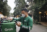 A delivery person dressed in green attire and a face mask is interacting with a mobile device while checking a matching green delivery bag. The background features a tree-lined street with buildings and vehicles, suggesting an urban setting.