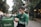 A delivery person dressed in green attire and a face mask is interacting with a mobile device while checking a matching green delivery bag. The background features a tree-lined street with buildings and vehicles, suggesting an urban setting.