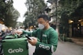 A delivery person dressed in green attire and a face mask is interacting with a mobile device while checking a matching green delivery bag. The background features a tree-lined street with buildings and vehicles, suggesting an urban setting.