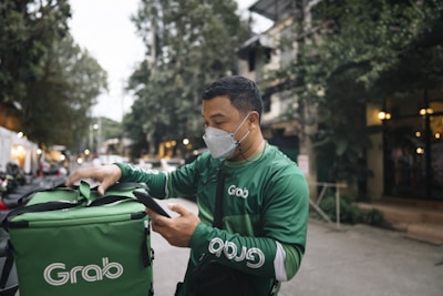 A delivery person dressed in green attire and a face mask is interacting with a mobile device while checking a matching green delivery bag. The background features a tree-lined street with buildings and vehicles, suggesting an urban setting.