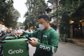 A delivery person dressed in green attire and a face mask is interacting with a mobile device while checking a matching green delivery bag. The background features a tree-lined street with buildings and vehicles, suggesting an urban setting.
