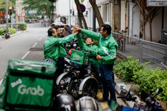 Several delivery riders wearing green Grab jackets are gathered on the sidewalk giving each other high-fives. They are standing next to their parked motorcycles and bicycles, and one of them holds a helmet. The setting appears to be an urban street lined with greenery and buildings.