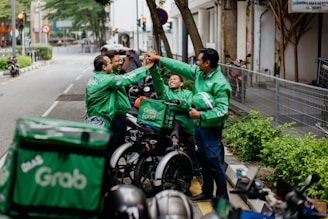 Several delivery riders wearing green Grab jackets are gathered on the sidewalk giving each other high-fives. They are standing next to their parked motorcycles and bicycles, and one of them holds a helmet. The setting appears to be an urban street lined with greenery and buildings.