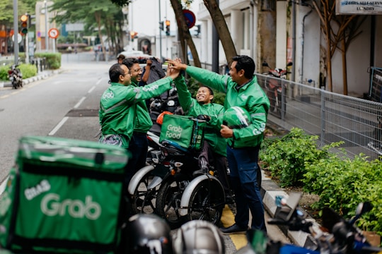 Several delivery riders wearing green Grab jackets are gathered on the sidewalk giving each other high-fives. They are standing next to their parked motorcycles and bicycles, and one of them holds a helmet. The setting appears to be an urban street lined with greenery and buildings.