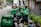 Several delivery riders wearing green Grab jackets are gathered on the sidewalk giving each other high-fives. They are standing next to their parked motorcycles and bicycles, and one of them holds a helmet. The setting appears to be an urban street lined with greenery and buildings.