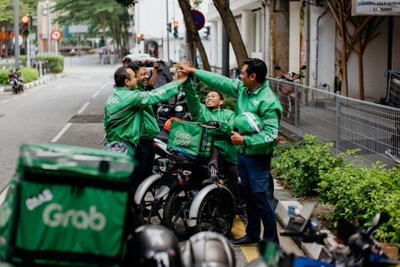 Several delivery riders wearing green Grab jackets are gathered on the sidewalk giving each other high-fives. They are standing next to their parked motorcycles and bicycles, and one of them holds a helmet. The setting appears to be an urban street lined with greenery and buildings.