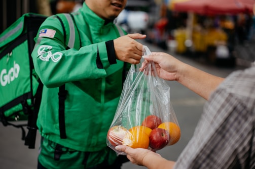 A delivery person in a green uniform is handing over a plastic bag containing fresh fruits, like oranges and apples, to another person. The interaction takes place outdoors, possibly at a market.