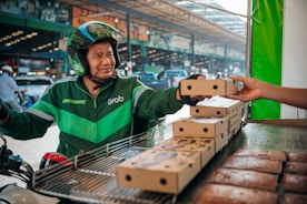 A delivery person wearing a green Grab uniform and helmet is on a motorcycle, accepting a boxed package from a hand reaching out towards them. The setting appears to be an outdoor market or street with blurred vehicles and buildings in the background.