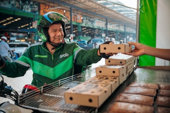A delivery person wearing a green Grab uniform and helmet is on a motorcycle, accepting a boxed package from a hand reaching out towards them. The setting appears to be an outdoor market or street with blurred vehicles and buildings in the background.