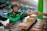 A delivery person wearing a green Grab uniform and helmet is on a motorcycle, accepting a boxed package from a hand reaching out towards them. The setting appears to be an outdoor market or street with blurred vehicles and buildings in the background.