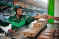 A delivery person wearing a green Grab uniform and helmet is on a motorcycle, accepting a boxed package from a hand reaching out towards them. The setting appears to be an outdoor market or street with blurred vehicles and buildings in the background.