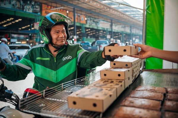 A delivery person wearing a green Grab uniform and helmet is on a motorcycle, accepting a boxed package from a hand reaching out towards them. The setting appears to be an outdoor market or street with blurred vehicles and buildings in the background.