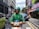 A delivery worker wearing a green uniform and helmet is riding a scooter on a city street. There are buildings and parked cars in the background, with some pedestrians visible on the sidewalk.