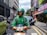 A delivery worker wearing a green uniform and helmet is riding a scooter on a city street. There are buildings and parked cars in the background, with some pedestrians visible on the sidewalk.