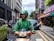 A delivery worker wearing a green uniform and helmet is riding a scooter on a city street. There are buildings and parked cars in the background, with some pedestrians visible on the sidewalk.