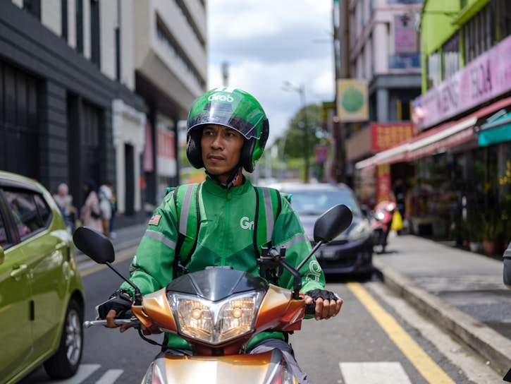 A delivery worker wearing a green uniform and helmet is riding a scooter on a city street. There are buildings and parked cars in the background, with some pedestrians visible on the sidewalk.