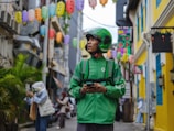 A delivery person wearing a green uniform and helmet stands in an alleyway surrounded by colorful lanterns hanging overhead. The street is lively with pedestrians, and buildings with bright yellow and blue accents line the narrow path.