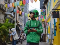 A delivery person wearing a green uniform and helmet stands in an alleyway surrounded by colorful lanterns hanging overhead. The street is lively with pedestrians, and buildings with bright yellow and blue accents line the narrow path.