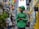 A delivery person wearing a green uniform and helmet stands in an alleyway surrounded by colorful lanterns hanging overhead. The street is lively with pedestrians, and buildings with bright yellow and blue accents line the narrow path.