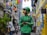 A delivery person wearing a green uniform and helmet stands in an alleyway surrounded by colorful lanterns hanging overhead. The street is lively with pedestrians, and buildings with bright yellow and blue accents line the narrow path.