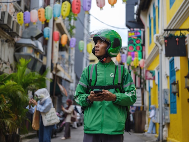 A delivery person wearing a green uniform and helmet stands in an alleyway surrounded by colorful lanterns hanging overhead. The street is lively with pedestrians, and buildings with bright yellow and blue accents line the narrow path.
