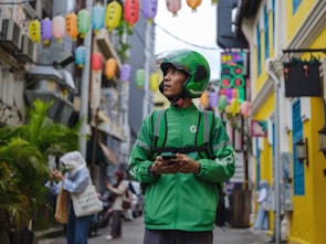 A delivery person wearing a green uniform and helmet stands in an alleyway surrounded by colorful lanterns hanging overhead. The street is lively with pedestrians, and buildings with bright yellow and blue accents line the narrow path.