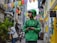 A delivery person wearing a green uniform and helmet stands in an alleyway surrounded by colorful lanterns hanging overhead. The street is lively with pedestrians, and buildings with bright yellow and blue accents line the narrow path.