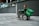 A person in a wheelchair equipped with a large green delivery bag labeled 'Grab' is on a gray tiled urban sidewalk. The person wears a green jacket and appears to be maneuvering the wheelchair near a modern building with glass railings and geometric walls.