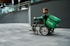 A person in a wheelchair equipped with a large green delivery bag labeled 'Grab' is on a gray tiled urban sidewalk. The person wears a green jacket and appears to be maneuvering the wheelchair near a modern building with glass railings and geometric walls.