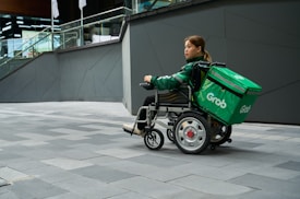 A person in a wheelchair equipped with a large green delivery bag labeled 'Grab' is on a gray tiled urban sidewalk. The person wears a green jacket and appears to be maneuvering the wheelchair near a modern building with glass railings and geometric walls.