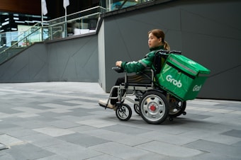 A person in a wheelchair equipped with a large green delivery bag labeled 'Grab' is on a gray tiled urban sidewalk. The person wears a green jacket and appears to be maneuvering the wheelchair near a modern building with glass railings and geometric walls.
