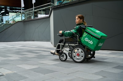 A person in a wheelchair equipped with a large green delivery bag labeled 'Grab' is on a gray tiled urban sidewalk. The person wears a green jacket and appears to be maneuvering the wheelchair near a modern building with glass railings and geometric walls.