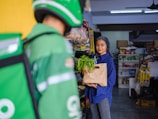A woman in a blue shirt and polka-dotted headscarf is holding a grocery bag with green vegetables. She is in a small store with shelves stocked with various products. A person wearing a green delivery uniform and helmet is in the foreground.
