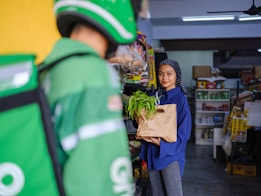 A woman in a blue shirt and polka-dotted headscarf is holding a grocery bag with green vegetables. She is in a small store with shelves stocked with various products. A person wearing a green delivery uniform and helmet is in the foreground.