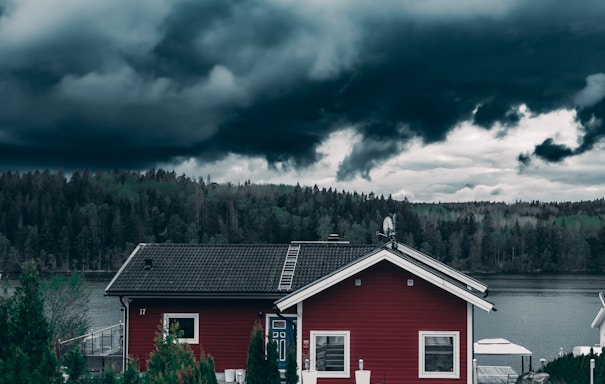 A small red house with white trim is set in a picturesque location by a body of water. Behind the house is a dense forest composed of diverse trees. Overhead, dark and dramatic storm clouds fill the sky, creating a stark contrast with the landscape.