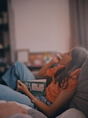 A person with long hair lying on a sofa, engrossed in reading a book with a colorful cover. They are wearing a bright orange shirt and blue jeans, and their wristwatch is visible. The background includes a blurred bookshelf and a window curtain, creating a cozy atmosphere.
