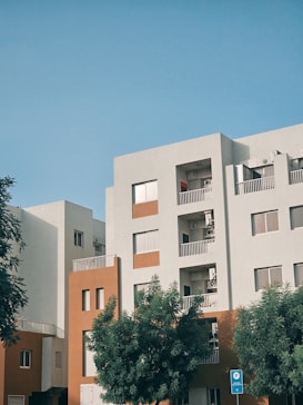A modern residential building with a minimalist design features multiple stories with balconies. The facade is a combination of white and burnt orange colors. In the foreground, there are lush green trees, and a clear blue sky forms the backdrop.