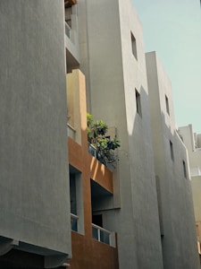 A modern residential building with a combination of smooth concrete surfaces and wooden textures. The architecture features clean lines and geometric shapes, with small windows integrated into the structure. There is a balcony with potted plants, adding a touch of greenery to the urban setting. The sunlight casts soft shadows across the facade, highlighting the building's textures.