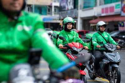 Three motorcyclists wearing green uniforms and helmets are gathered, each on a motorcycle. The central rider is on a red motorcycle, flanked by others on dark-colored bikes. Buildings and parked cars appear in the background.