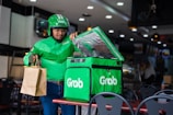 A delivery person wearing a green jacket and helmet holds a brown paper bag while standing beside a green insulated delivery bag with the logo 'Grab' on it. The setting appears to be inside a restaurant or food establishment with tables and chairs.