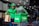 A delivery person wearing a green jacket and helmet holds a brown paper bag while standing beside a green insulated delivery bag with the logo 'Grab' on it. The setting appears to be inside a restaurant or food establishment with tables and chairs.
