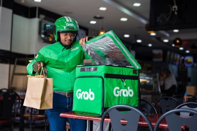 A delivery person wearing a green jacket and helmet holds a brown paper bag while standing beside a green insulated delivery bag with the logo 'Grab' on it. The setting appears to be inside a restaurant or food establishment with tables and chairs.