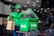 A delivery person wearing a green jacket and helmet holds a brown paper bag while standing beside a green insulated delivery bag with the logo 'Grab' on it. The setting appears to be inside a restaurant or food establishment with tables and chairs.